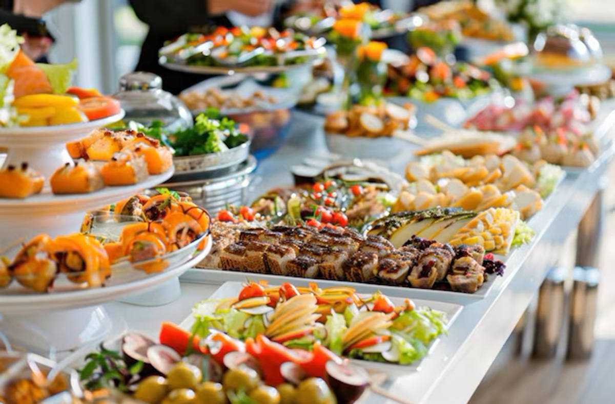 buffet food Buffet spread featuring a variety of hot and cold dishes in stainless steel trays, including grilled meats, rice, pasta, and vegetables, with serving utensils placed in each tray.