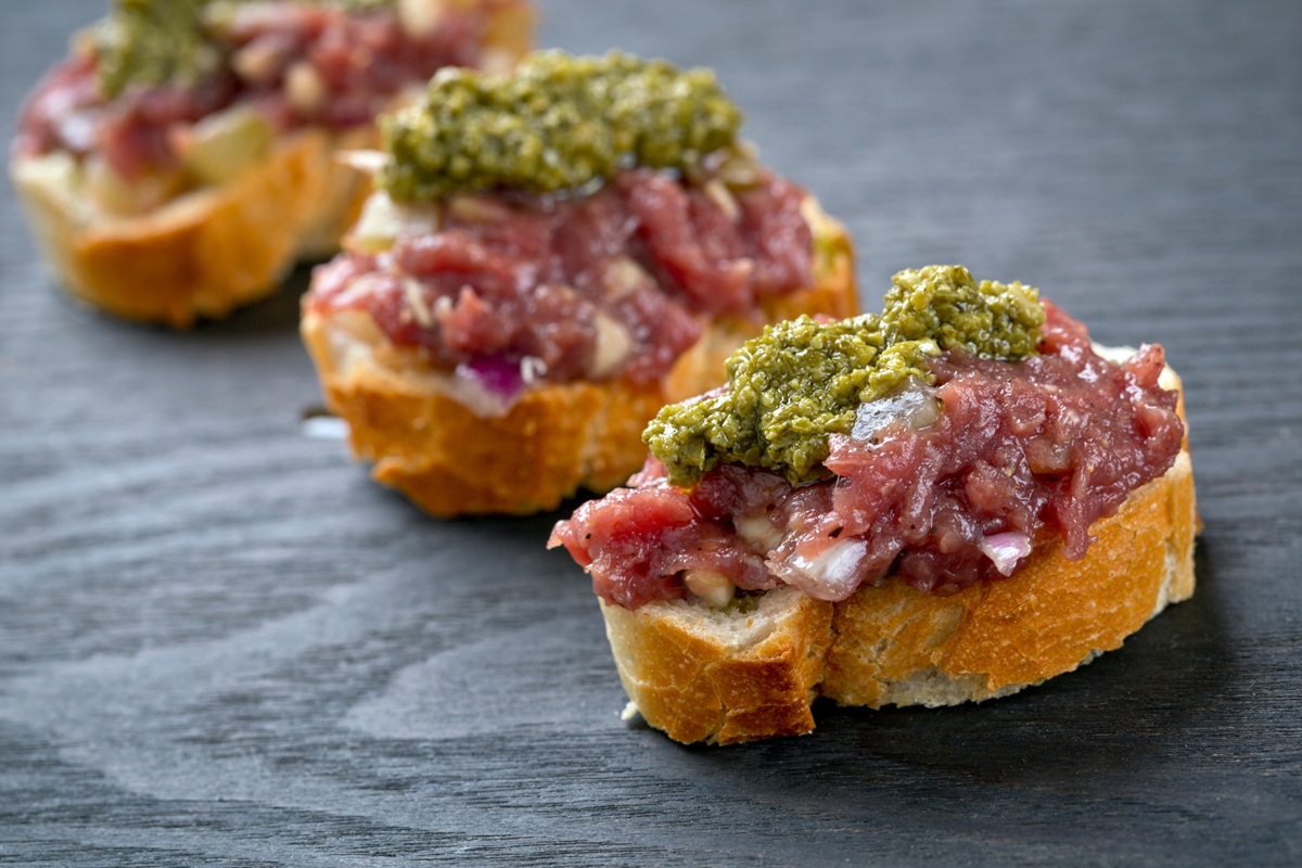 pics - 2 Close-up of three pieces of toasted baguette topped with beef tartare and a dollop of green pesto, arranged in a diagonal line on a dark slate surface.