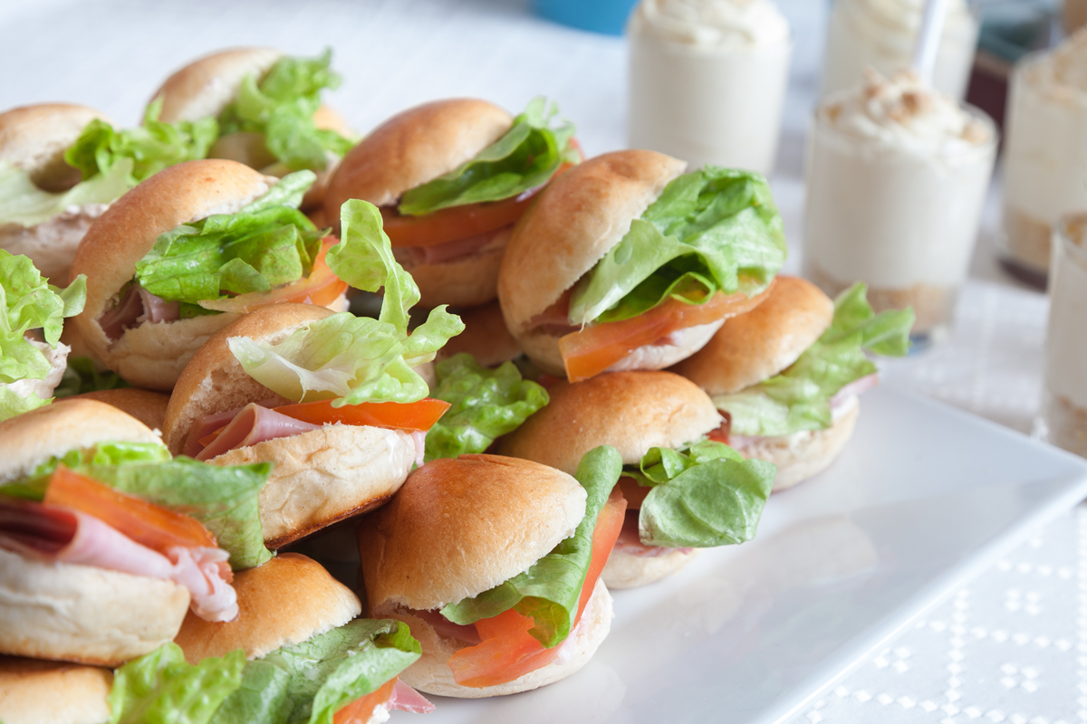 Tray of fresh mini sandwiches filled with lettuce, tomato, and sliced deli meat, served on a white tablecloth with dessert cups in the background.